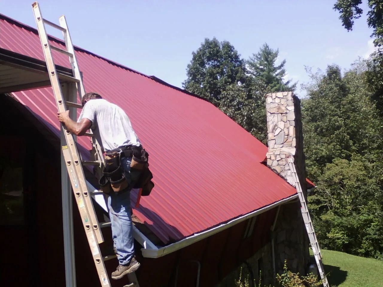 Roofer on a ladder beside a red metal roof and stone chimney — Soffit & Fascia Repair by Roofing Brothers in Rowland